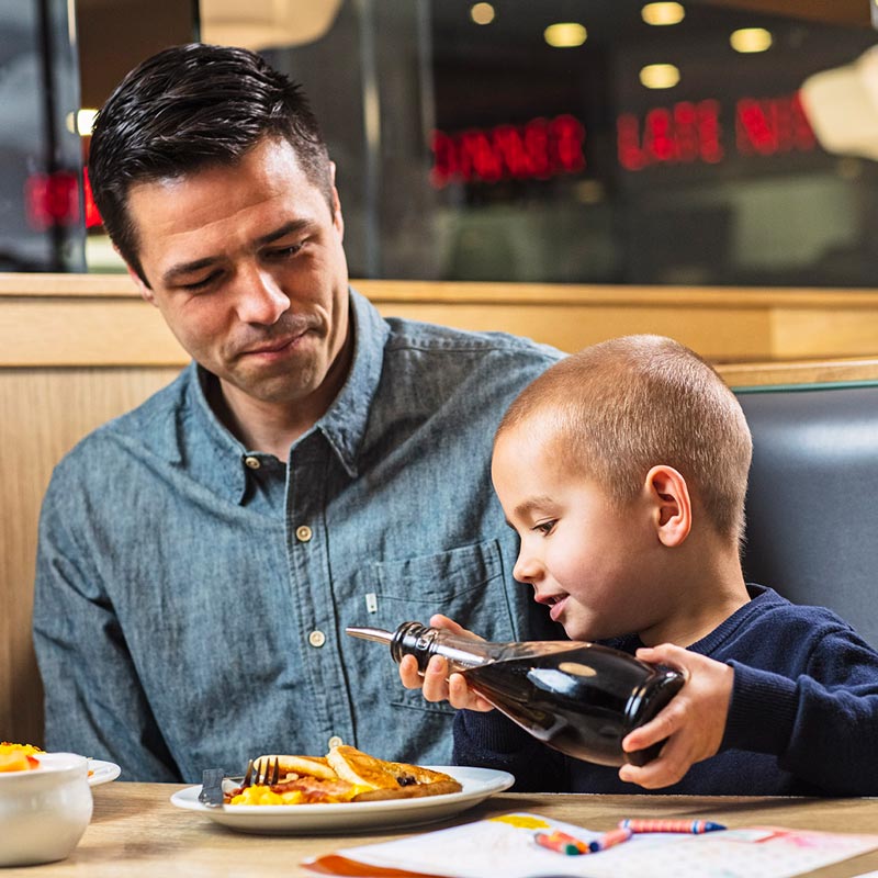 Father sitting with son pouring syrup onto a plate of pancakes